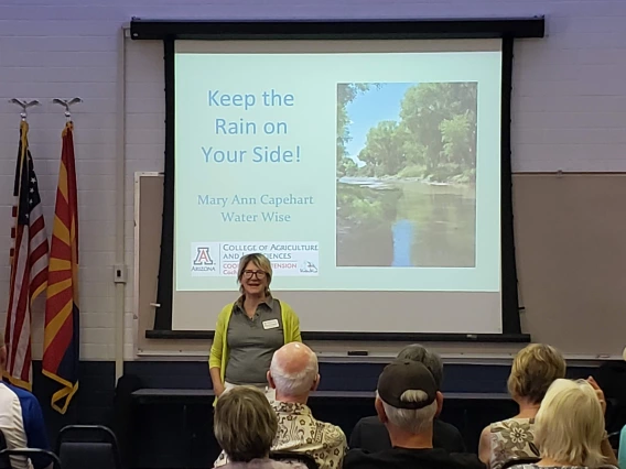 A light skinned woman with short blond hair wearing glasses stands in front of a projection of a slide show presentation entitled Keep Rain On Your Side