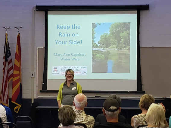 A light skinned woman with short blond hair wearing glasses stands in front of a projection of a slide show presentation entitled Keep Rain On Your Side