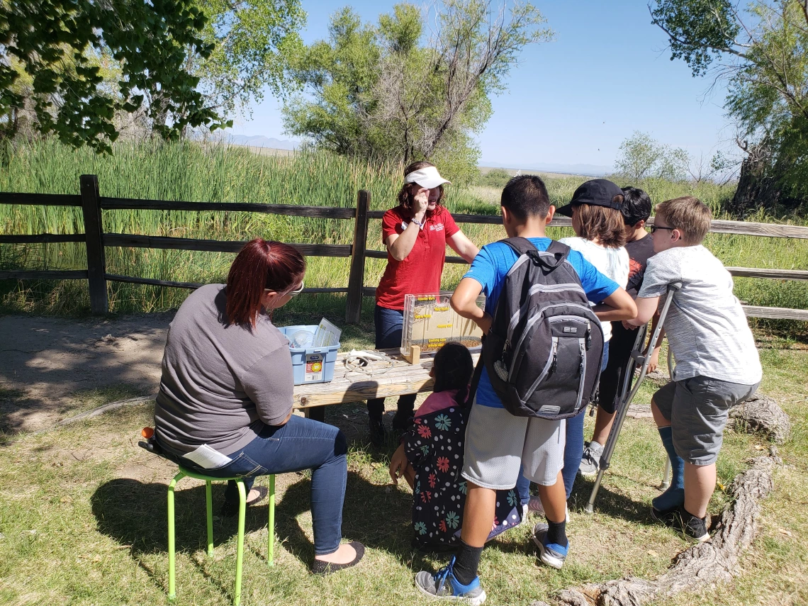 A Water Wise Youth Educator stands in nature instructing a group of six children with a groundwater flow model