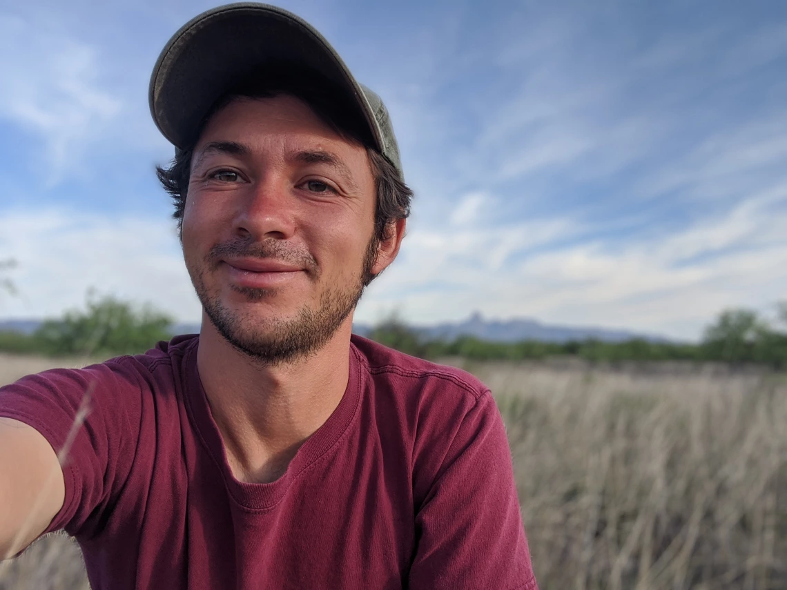 Trevor Lauber, a light skinned man with short dark hair wears a red shirt with a blue sky and grass in the background.
