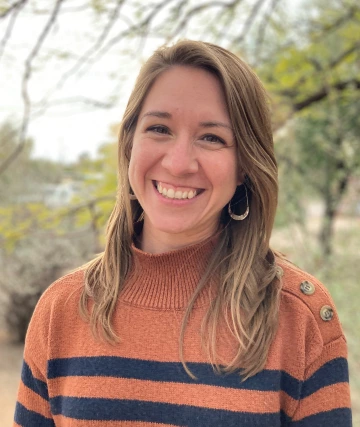 A light skinned woman with long blond hair smiles in a wooded area in an orange turtleneck sweater with navy stripes.