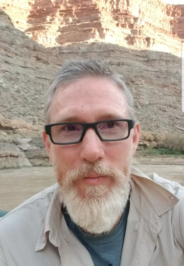 A light skinned man with gray hair and a gray beard and mustache stands in front of a rocky canyon with water below. He is wearing black glasses and a tan shirt.